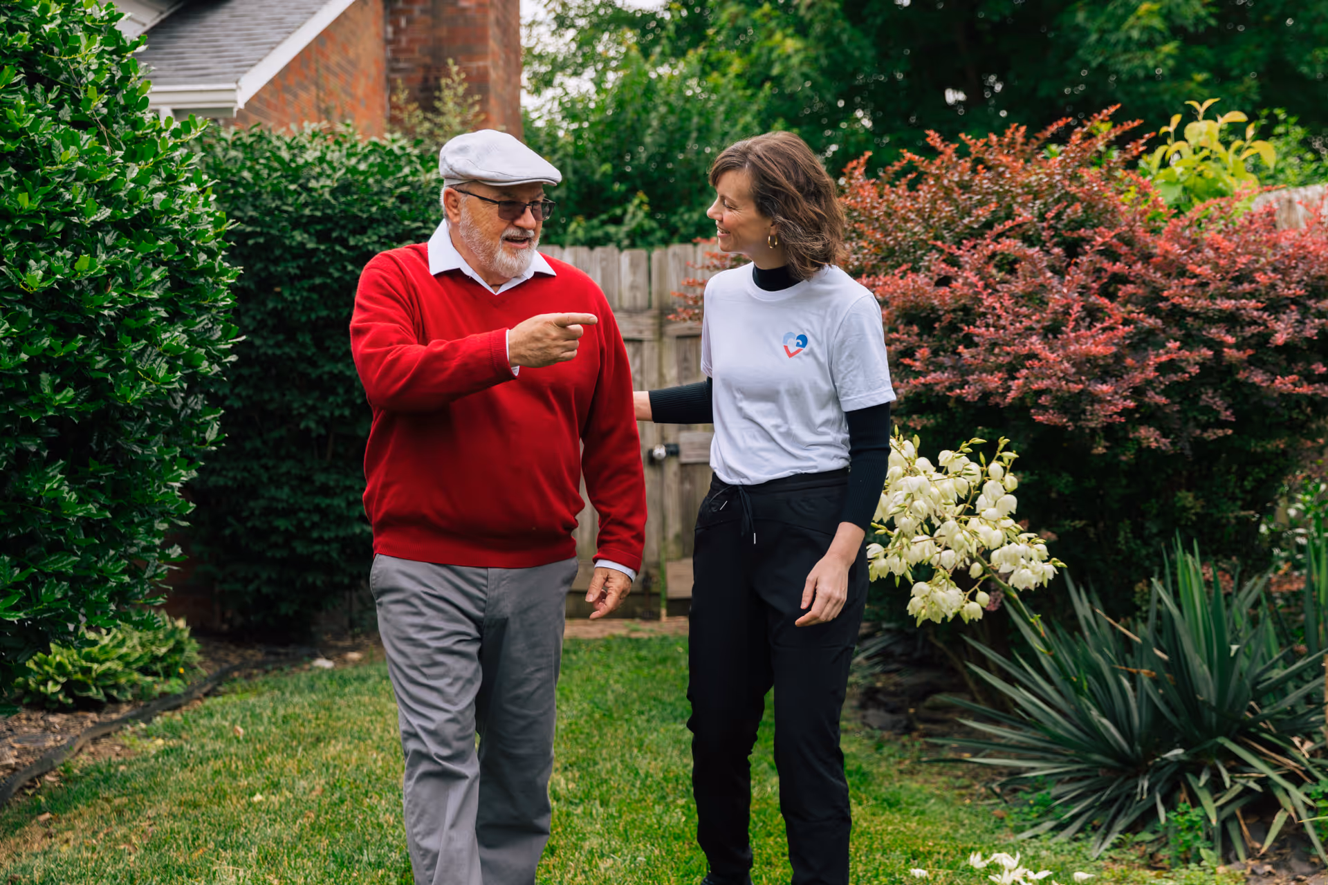 An older man and a caregiver converse in a well-kept backyard garden.