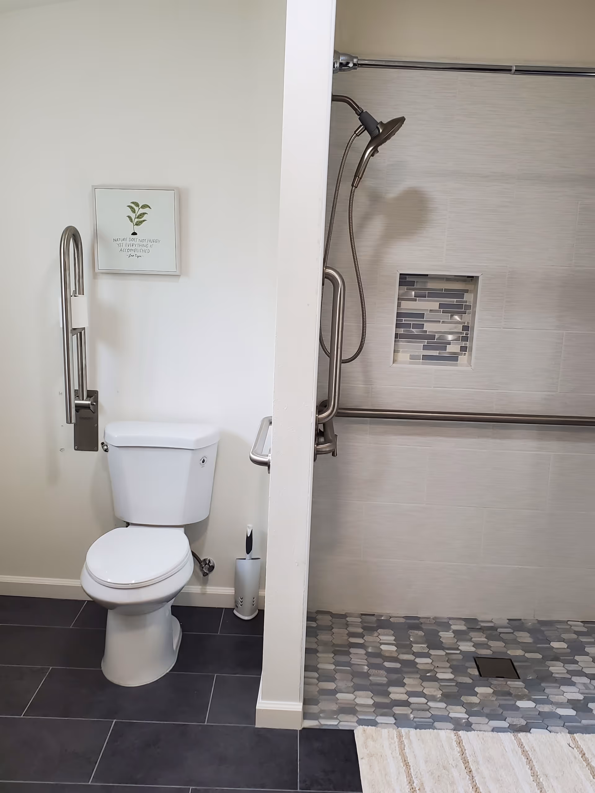 Accessible bathroom featuring toilet with grab bars, handheld shower, and tiled floor.