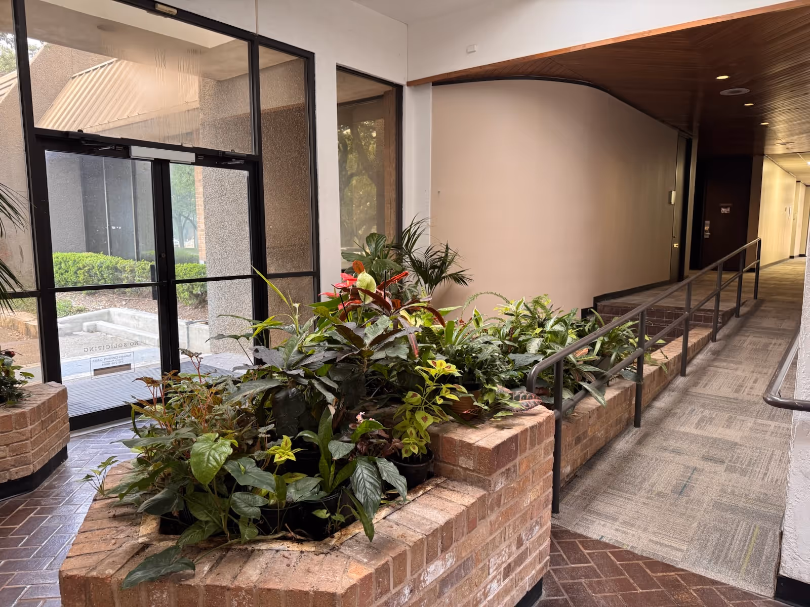Indoor hallway with brick planters and lush plants along a wheelchair accessible ramp.
