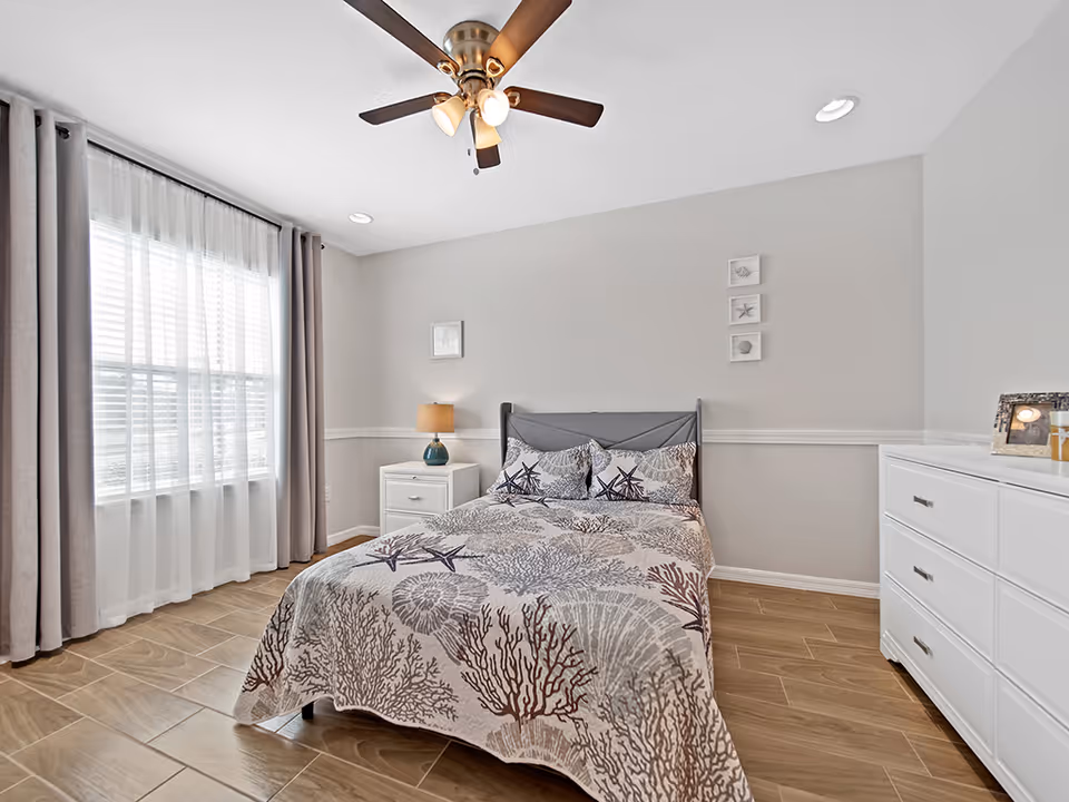 Bright bedroom featuring bed, nightstand, dresser, ceiling fan, and large window with curtains.