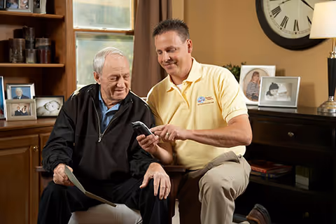 Caregiver sits with elderly man, showing him something on a handheld device in a living room.
