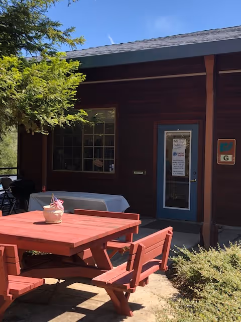Outdoor seating area with red picnic tables beside a brown building and greenery.