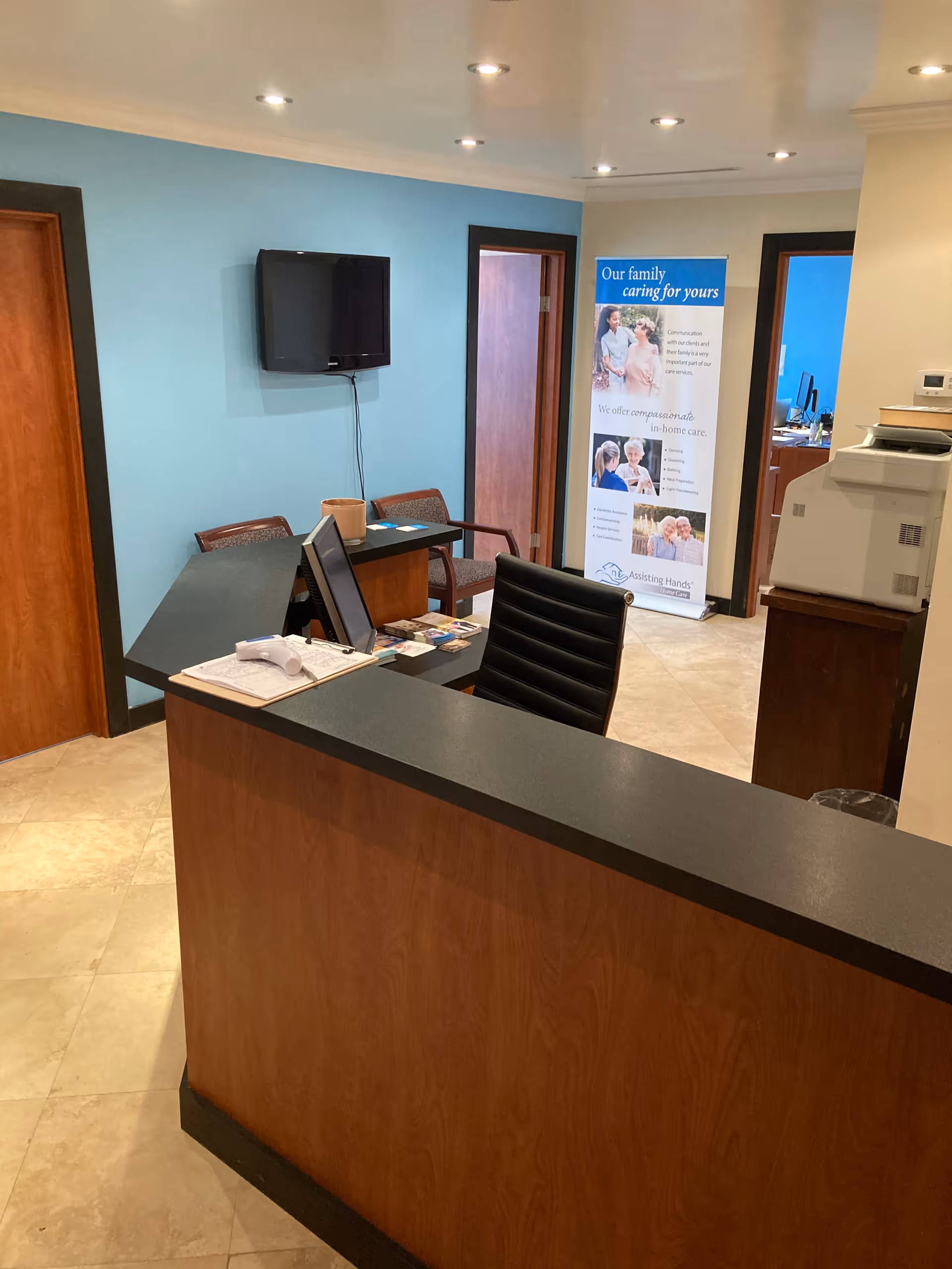 Reception area with curved wooden front desk, blue wall, TV, banner, and chairs.