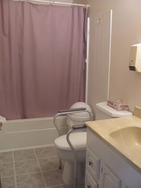 Bathroom in a senior living facility with purple shower curtain, toilet with bars, and sink.