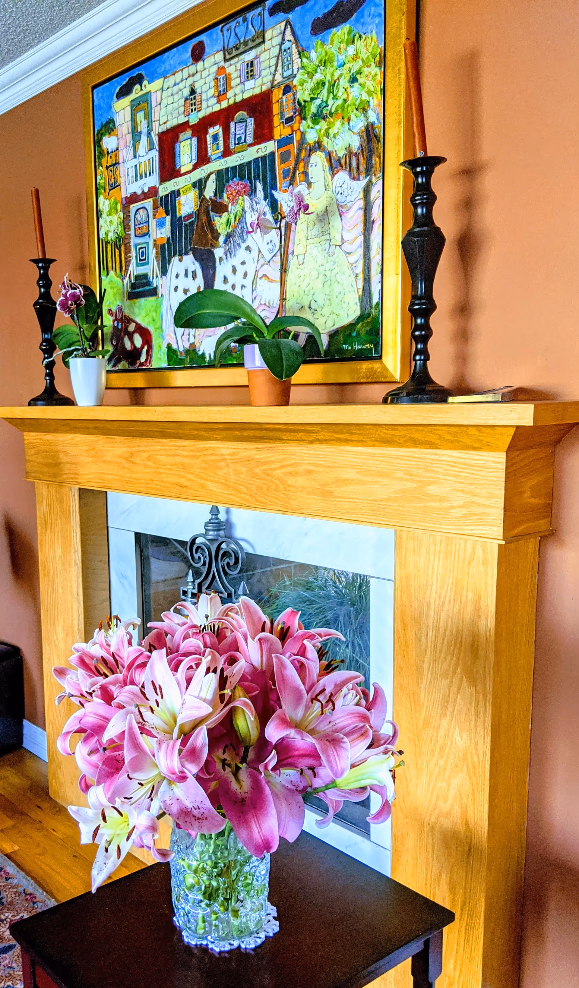 Bright living room with a wooden fireplace mantel, pink lilies bouquet, and a colorful painting above.