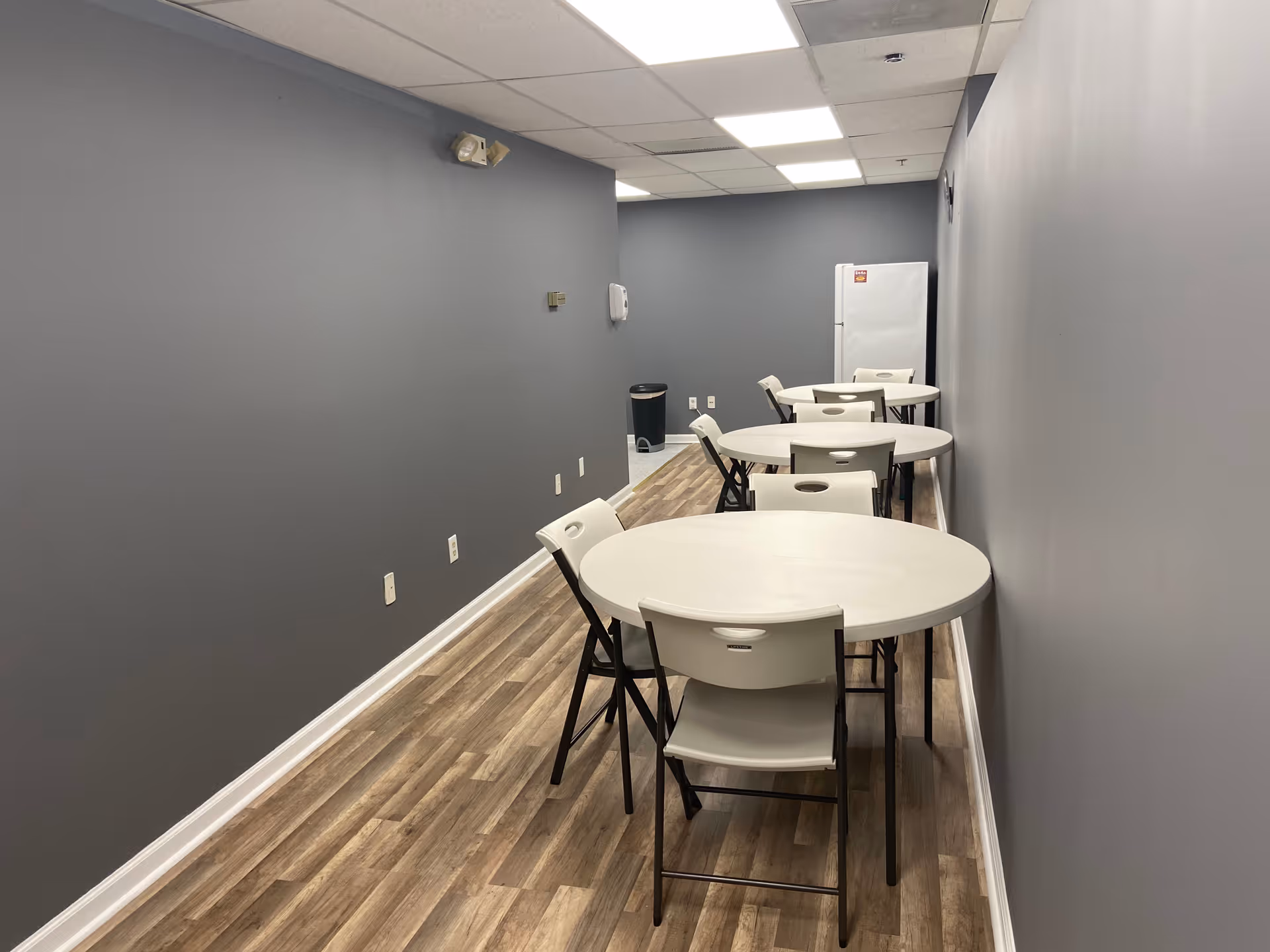 Narrow dining area with round tables and white chairs along gray walls.