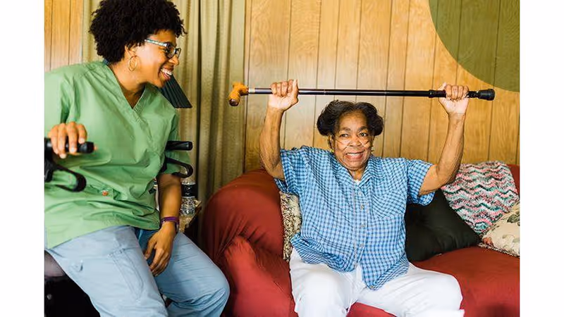 Caregiver in green scrubs assists elderly woman with overhead arm exercise indoors.