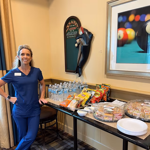 Caregiver in blue scrubs stands beside a snack and water table.