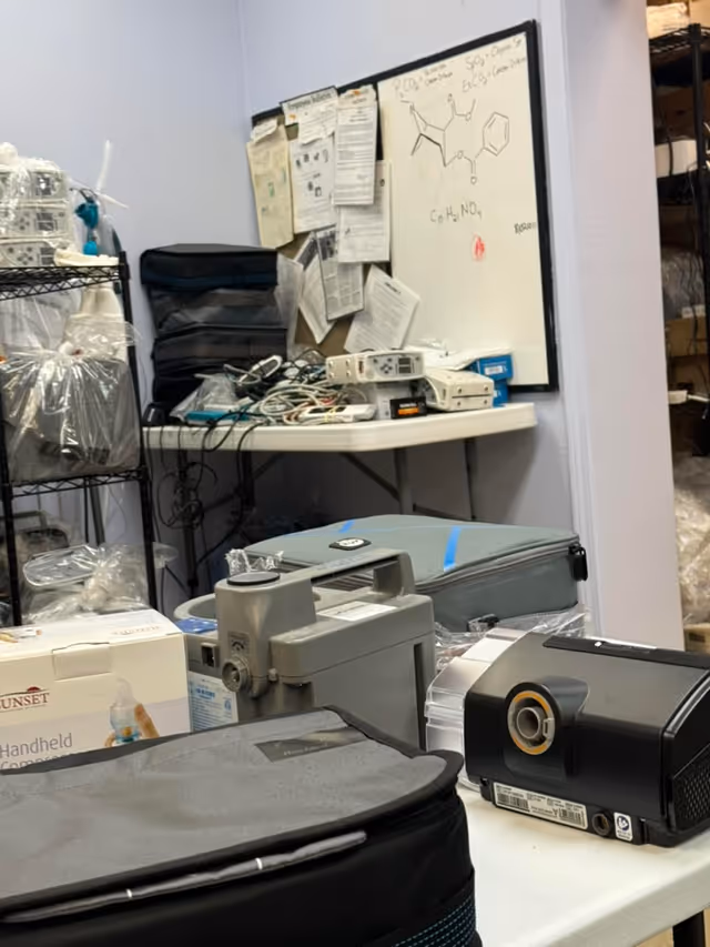 Cluttered storage room with medical devices, cables, boxes, and a whiteboard in LINCARE facility.