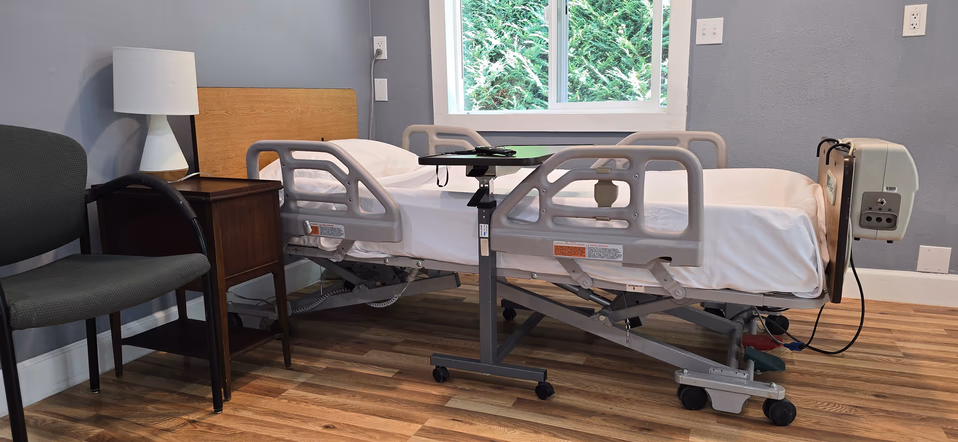 Hospital-style patient bed with rails, nightstand, lamp, and window in a quiet room.