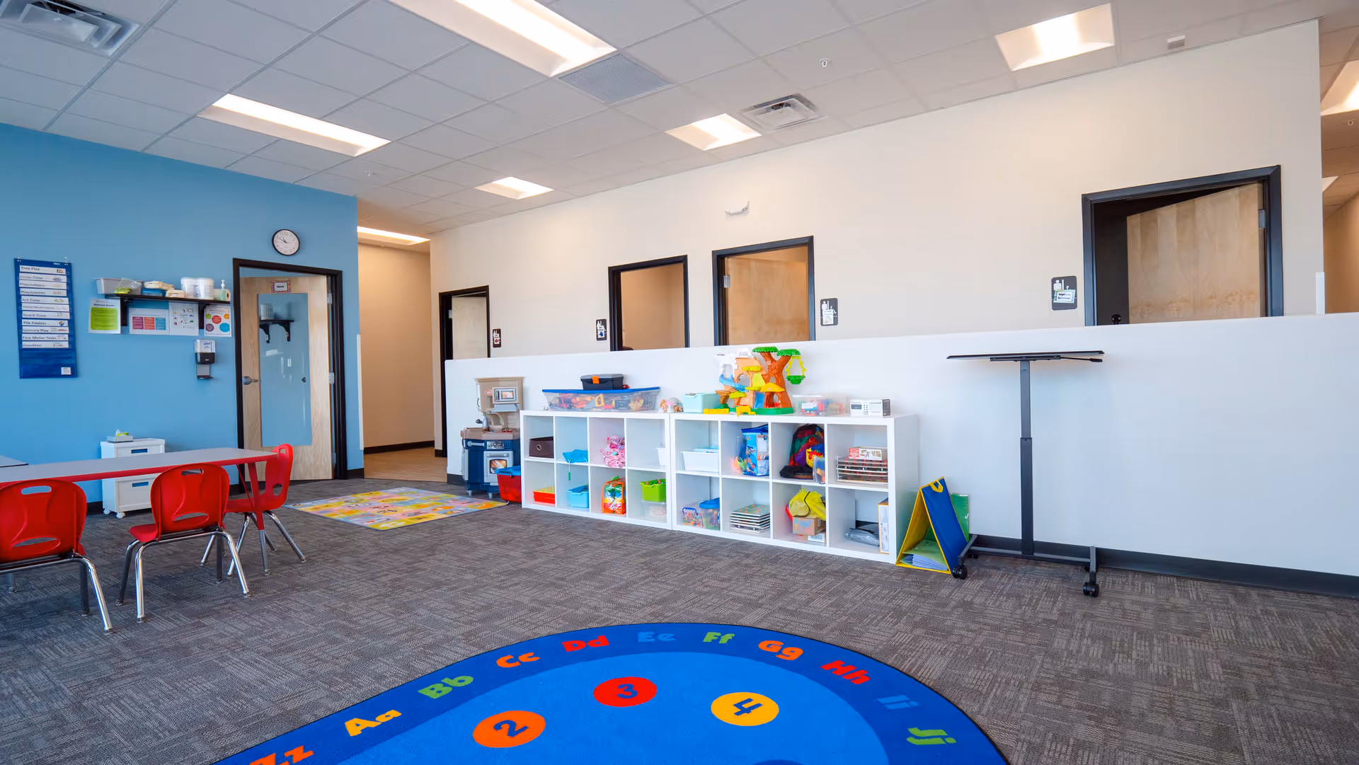 Bright children's playroom with toy shelves, tables, and a colorful circular rug.