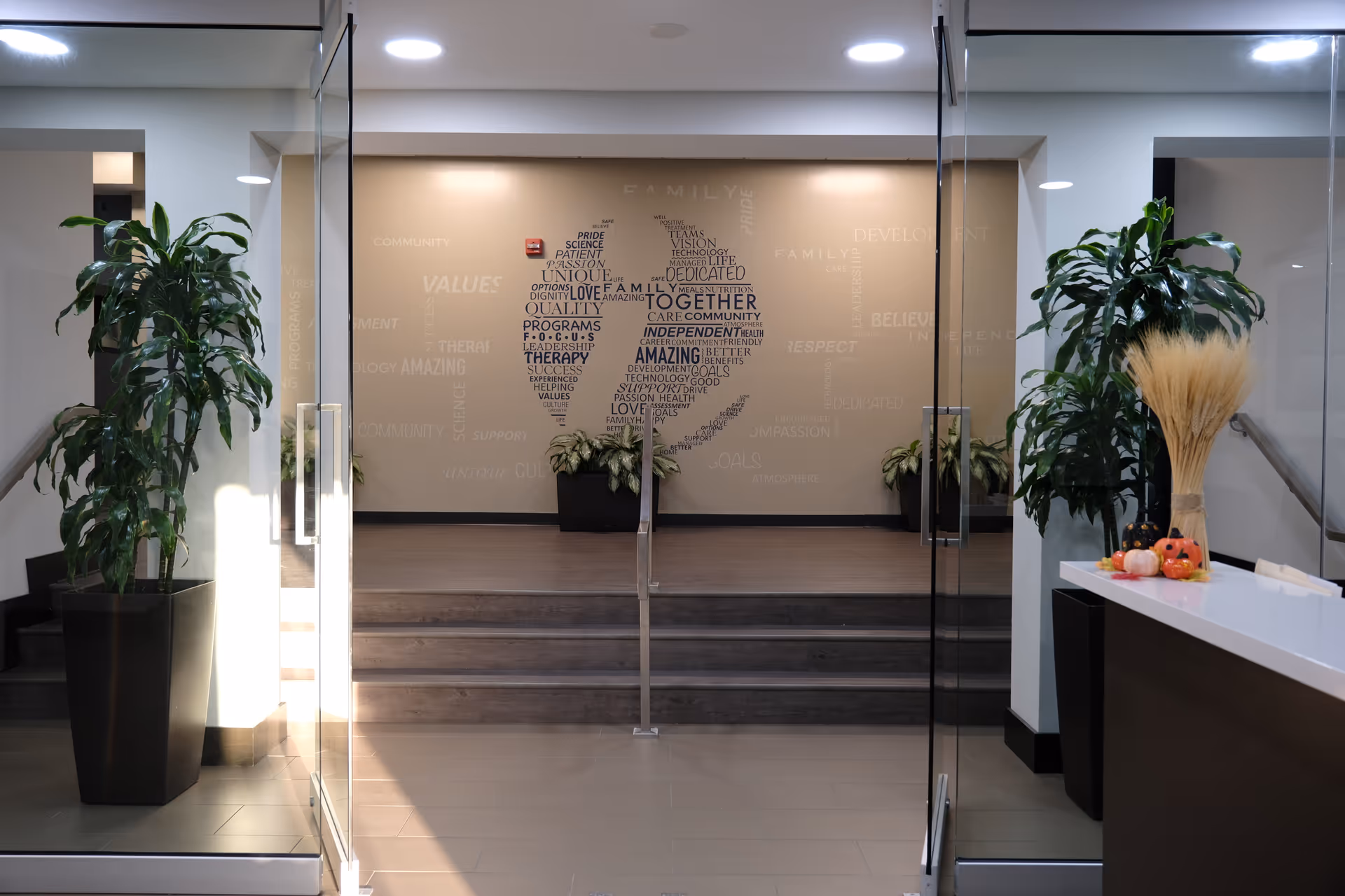 Modern facility lobby with glass doors, potted plants, stairs, and wall art.