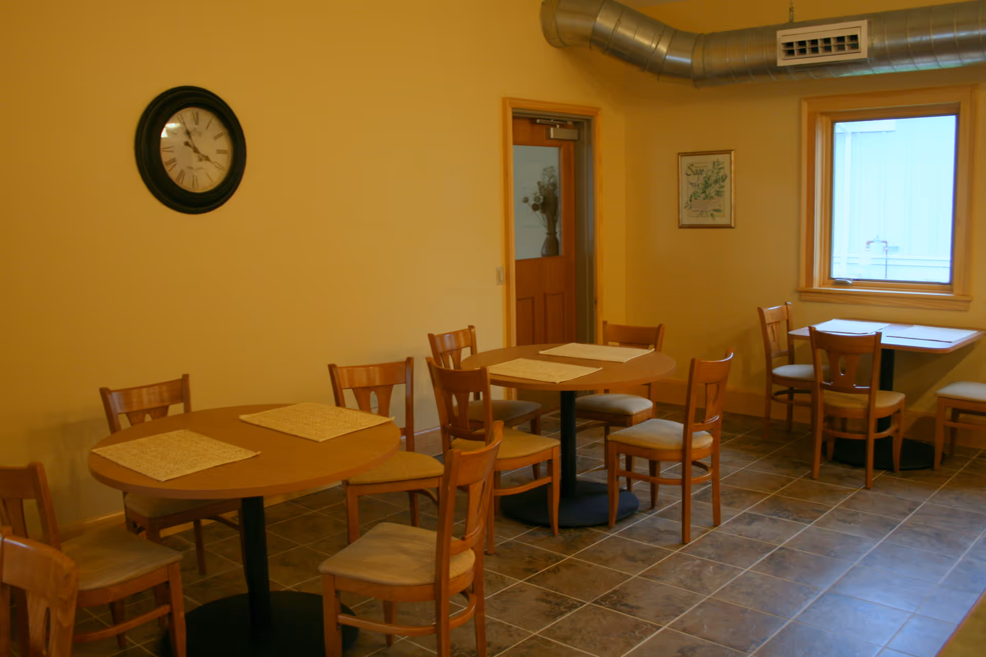 Warm yellow dining room with round wooden tables and matching chairs in a senior living facility.