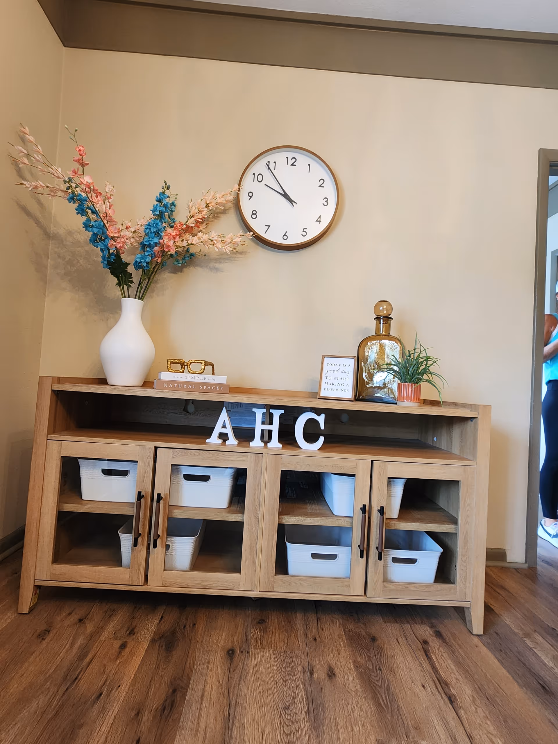 Wooden console with baskets, a vase of colorful flowers, and a wall clock.