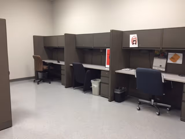 Row of gray cubicles with desks, chairs, and trash bins in an office area.