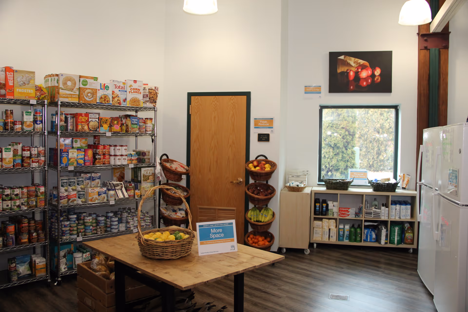 A compact kitchen/pantry area with grocery shelves, a table of fruit, and a refrigerator.