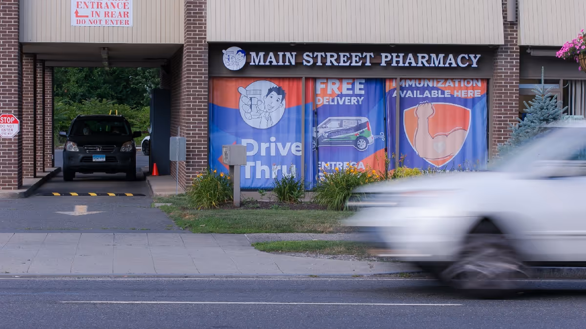 Brick storefront with Main Street Pharmacy sign, drive-thru window banners, motion-blurred car passing.
