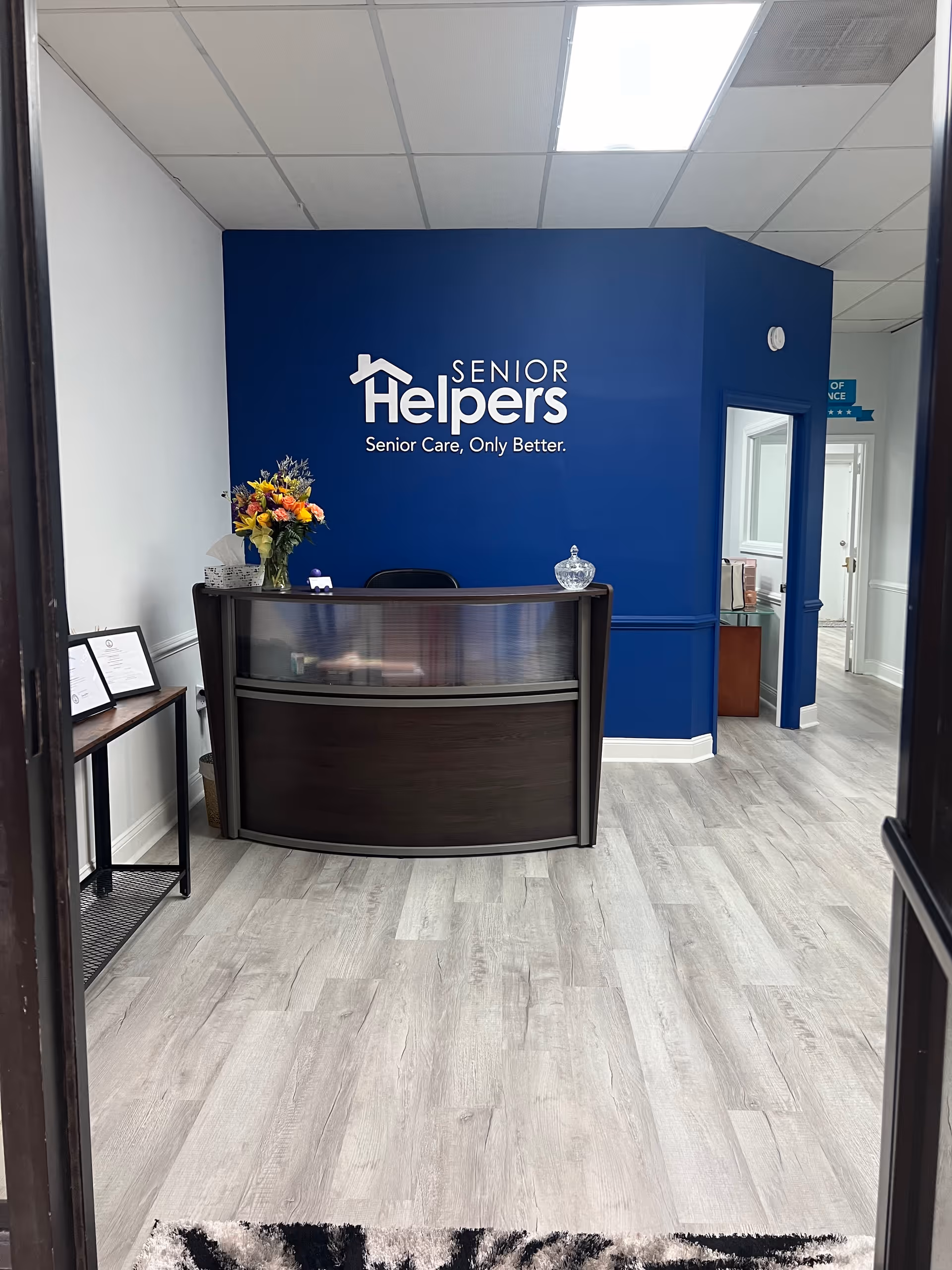 Reception desk area with blue wall branding and Senior Helpers logo, flowers, and paperwork.