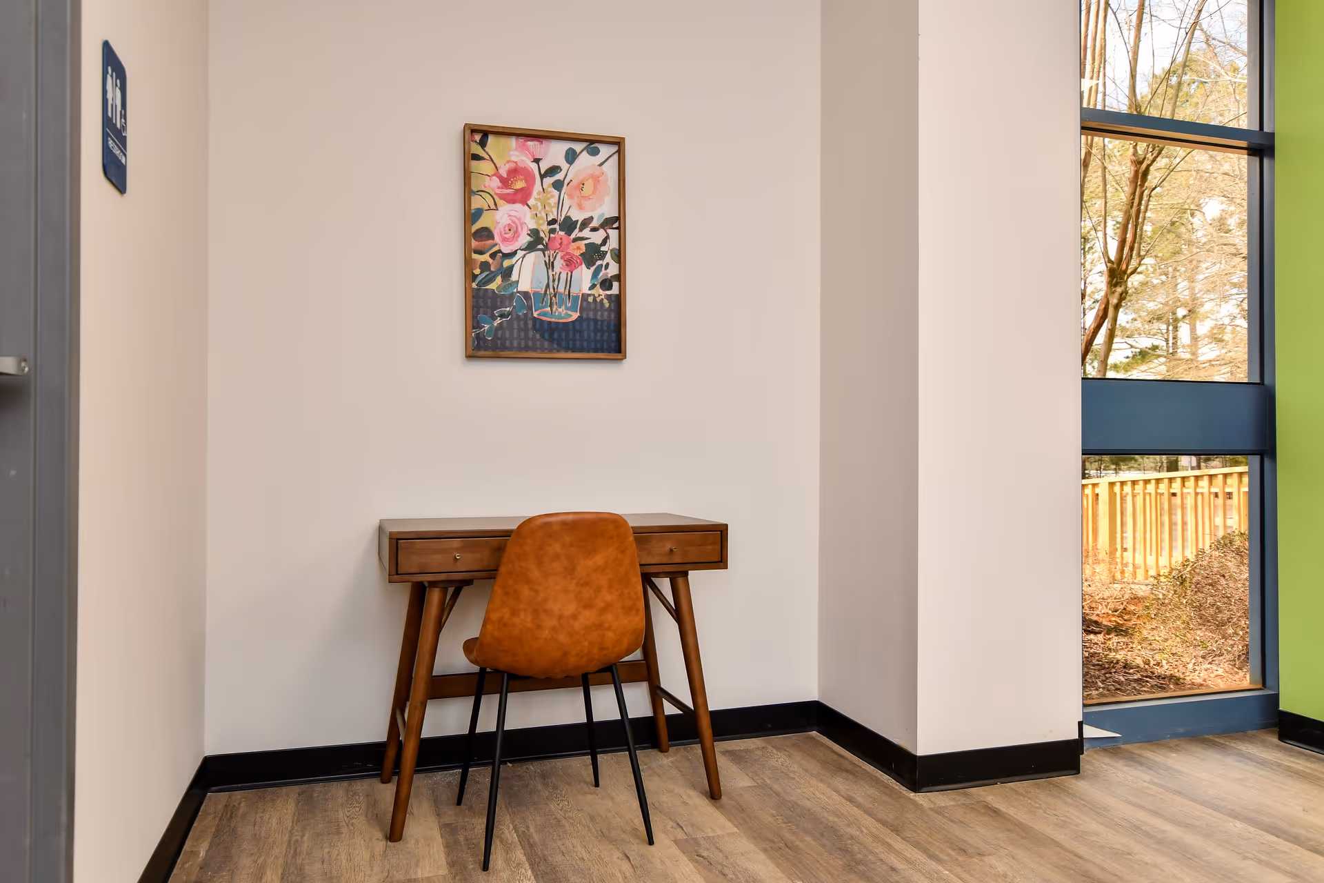 A wooden desk and orange chair sit in a corner under a floral painting.