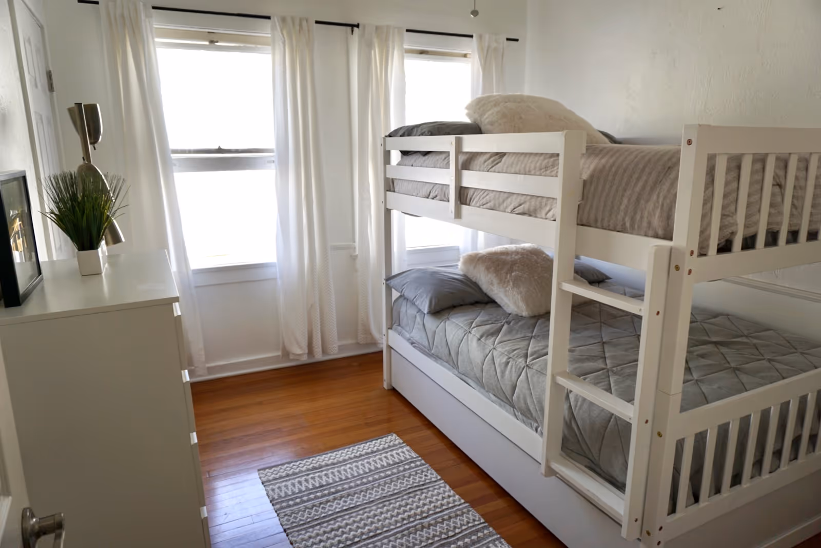 Bright bedroom with a white bunk bed, dresser, plant, and windows with curtains.