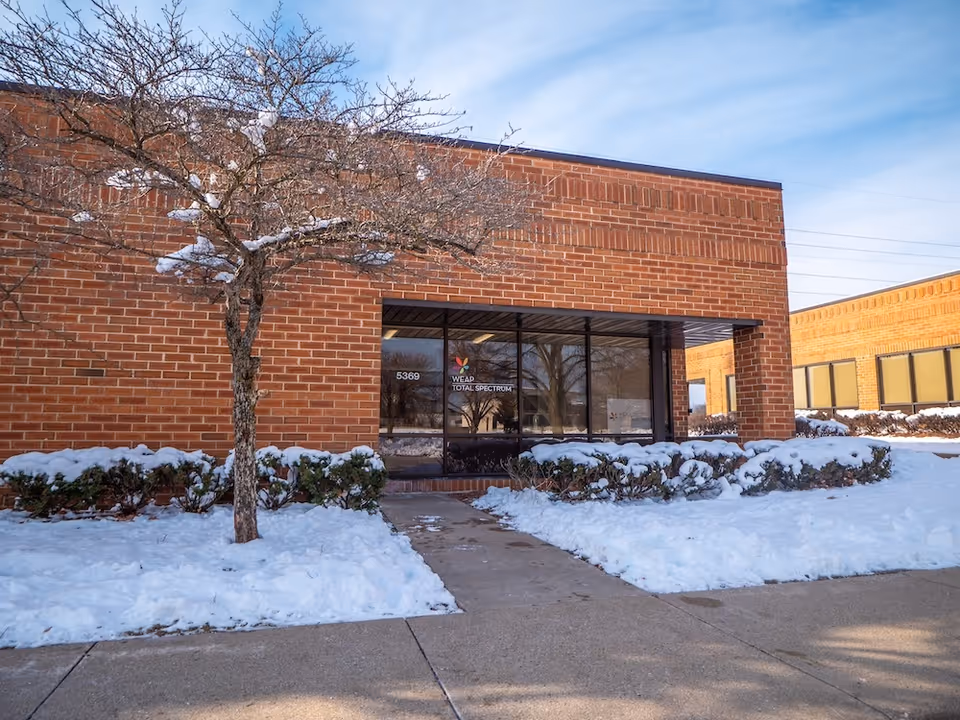 Red brick building entrance with glass doors, snow-covered landscaping, and a tree.