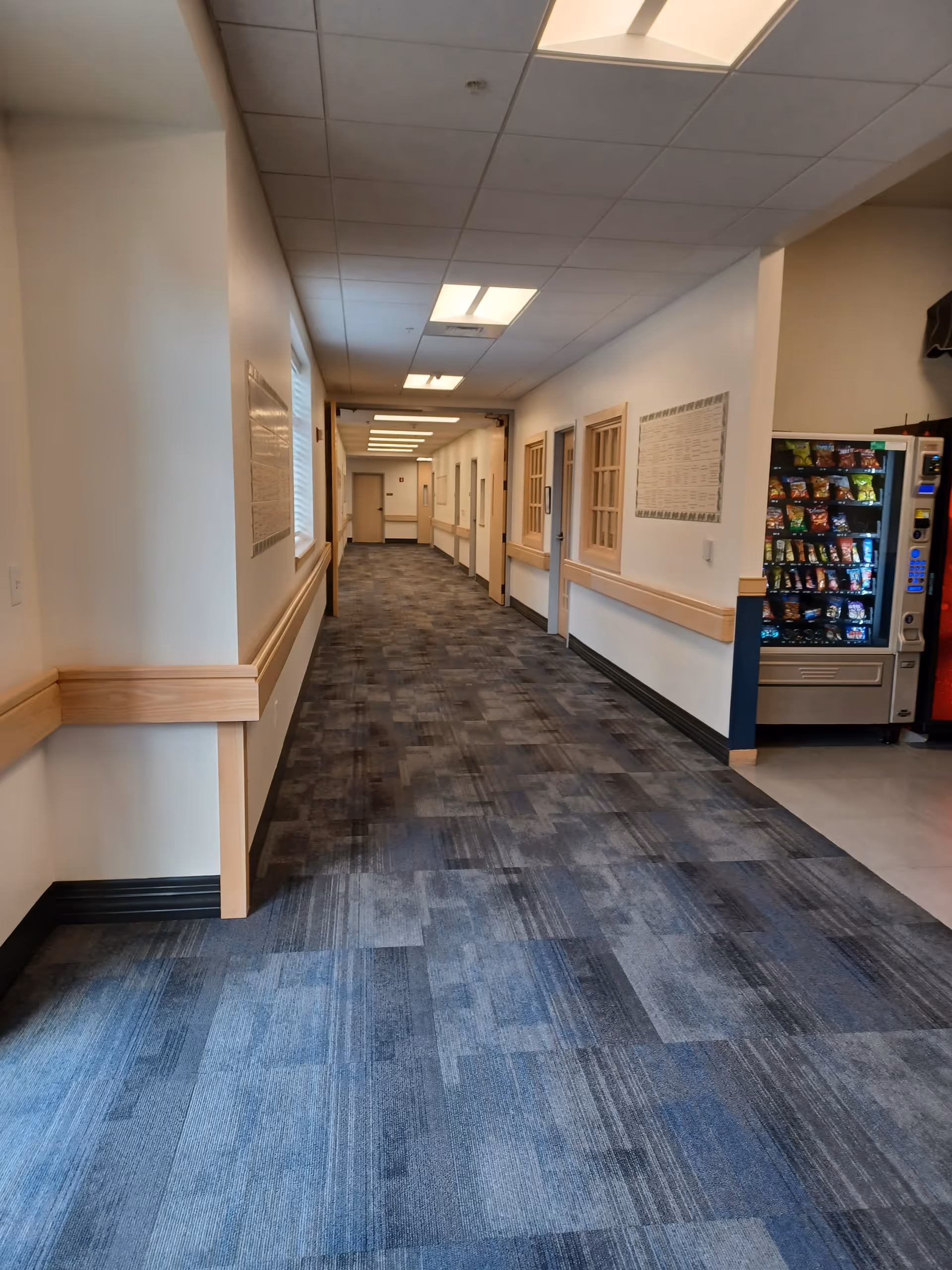 Long hallway in a senior living facility with doors, a vending machine, and carpet.