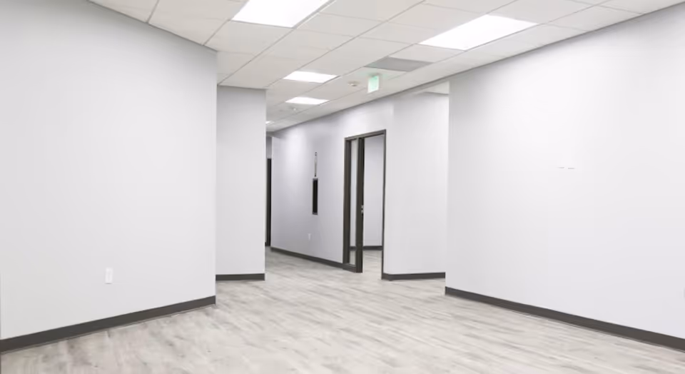 Long empty hallway with white walls, gray baseboards, and overhead fluorescent lighting.