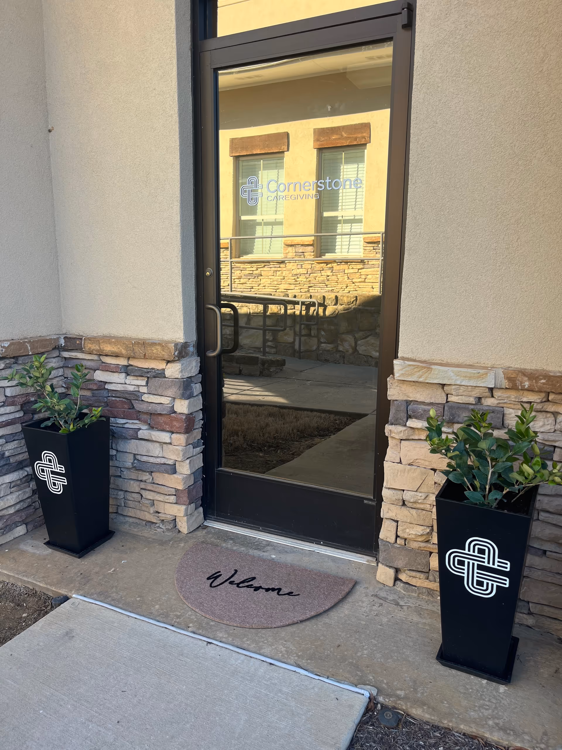 Exterior entrance to Cornerstone Caregiving with glass door, welcome mat, and potted plants.