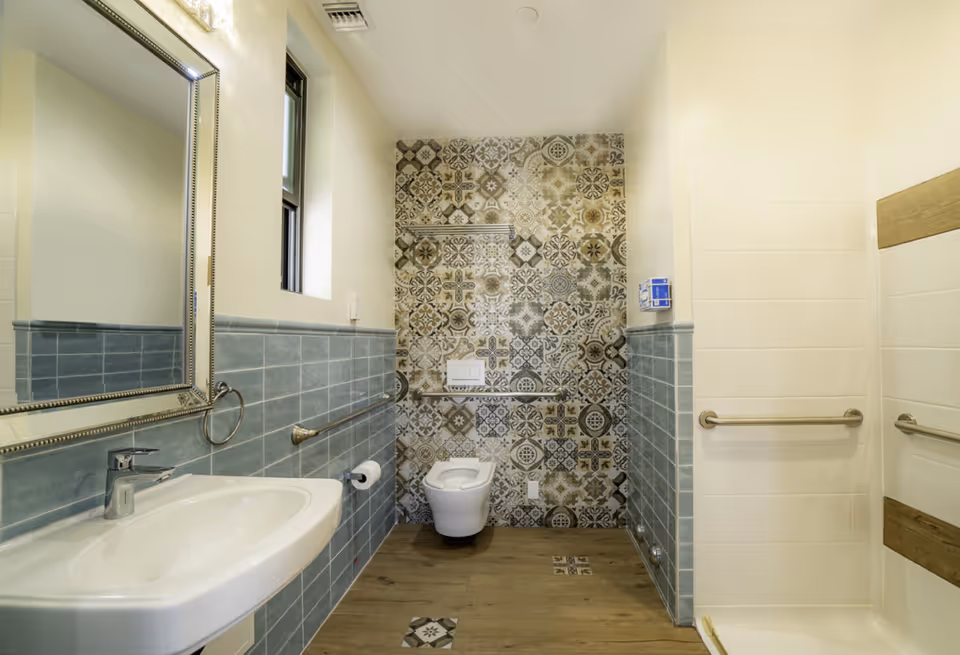 Senior living bathroom with sink, mirror, toilet, grab bars, and patterned tiled wall.