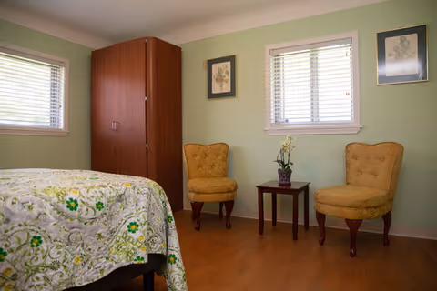 Light green bedroom featuring a bed, wardrobe, two yellow chairs, and a small table.