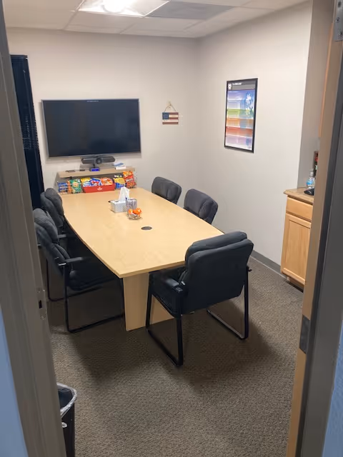A small conference room with a long wooden table, chairs, and a wall-mounted TV.
