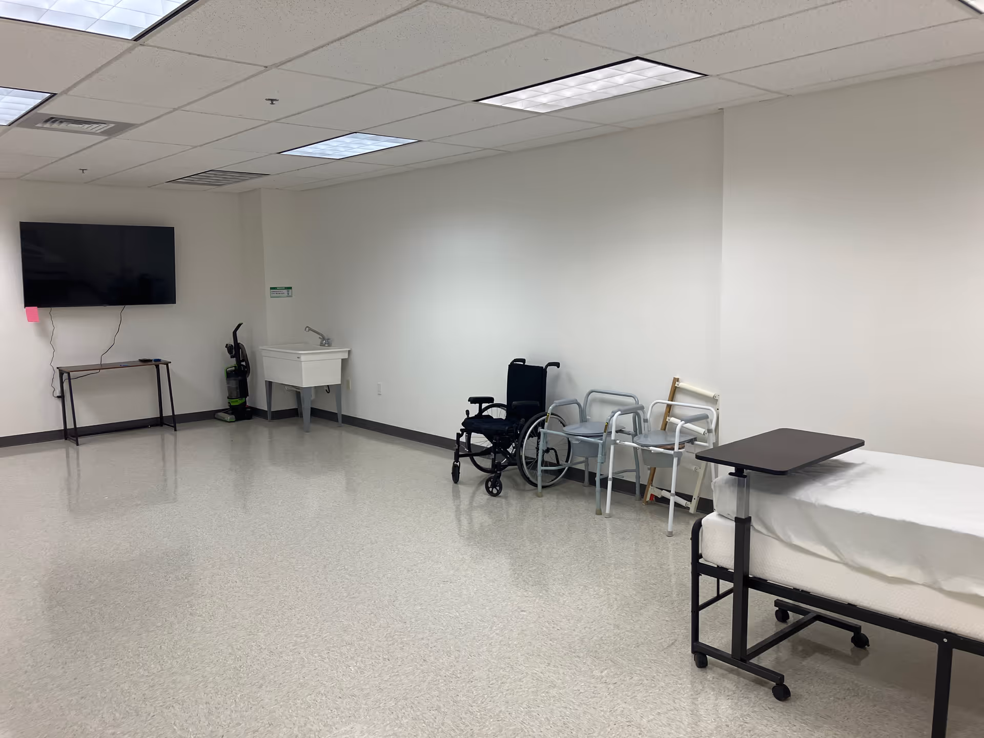 A spacious senior care room with a bed, wheelchair, walkers, sink, and wall TV.
