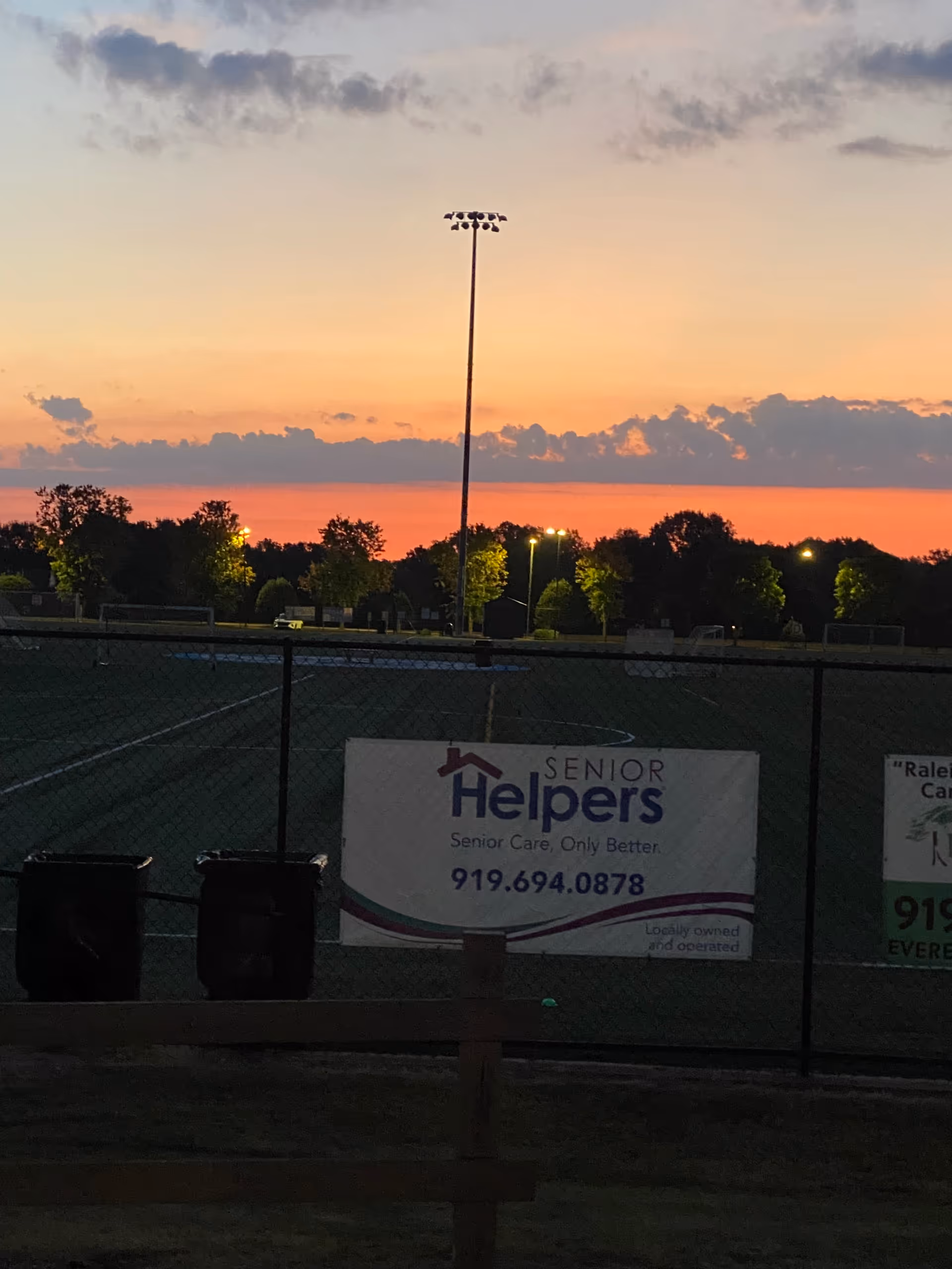Sunset over a fenced outdoor sports field with a Senior Helpers banner.