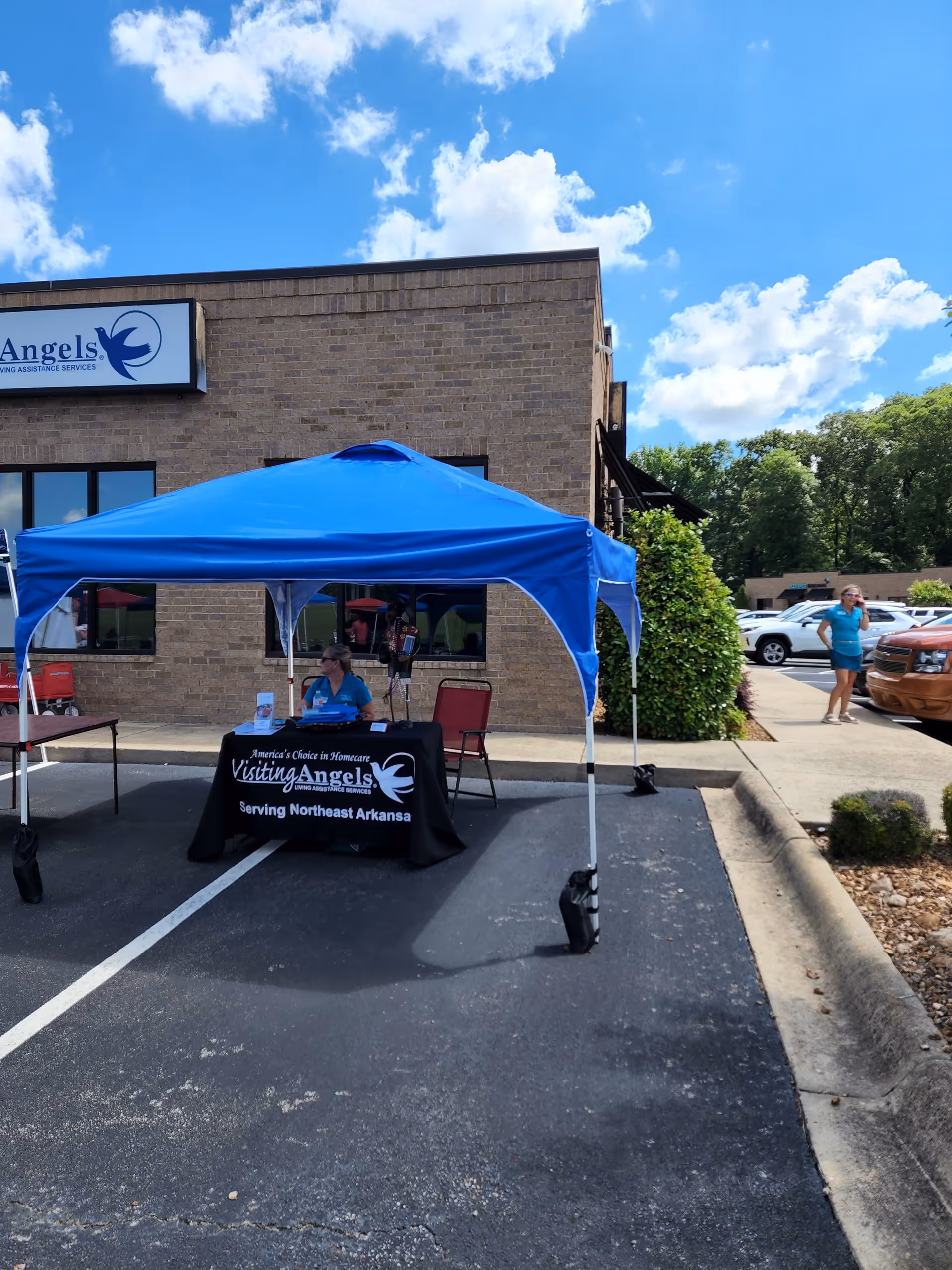 Staff member sits at a table under a blue canopy outside a brick senior care facility.