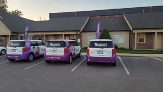 Row of purple Senior Helpers vans parked in front of a single story senior care facility.