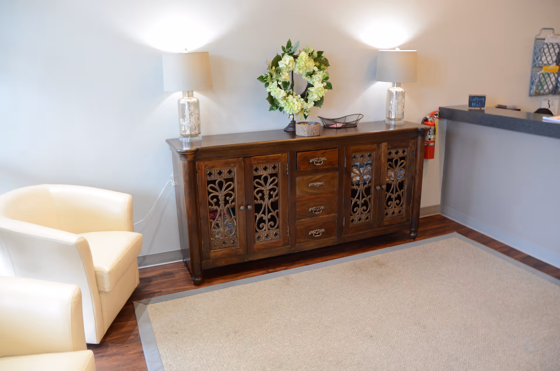 Reception area featuring a wooden cabinet, lamps, wreath, beige chairs, and a counter.
