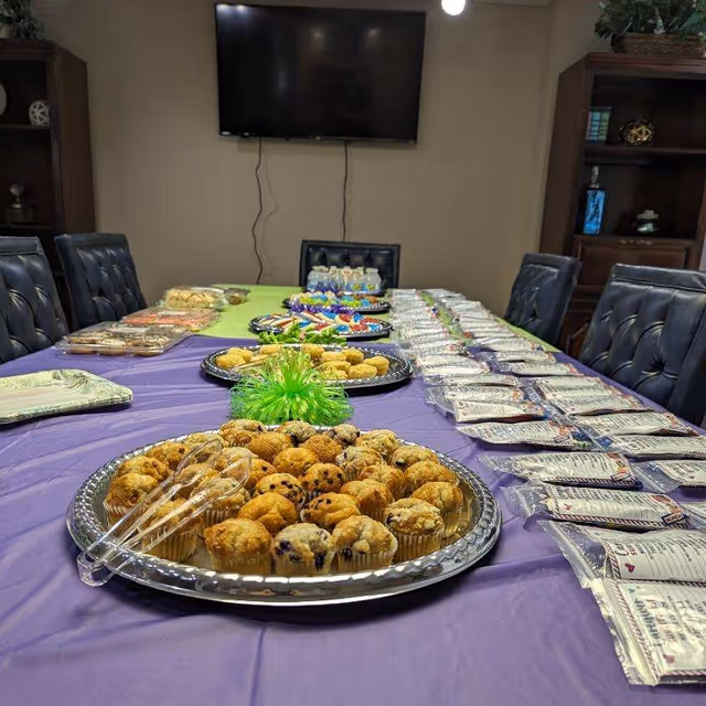A long table with purple tablecloth, pastries, and party supplies in a meeting room.