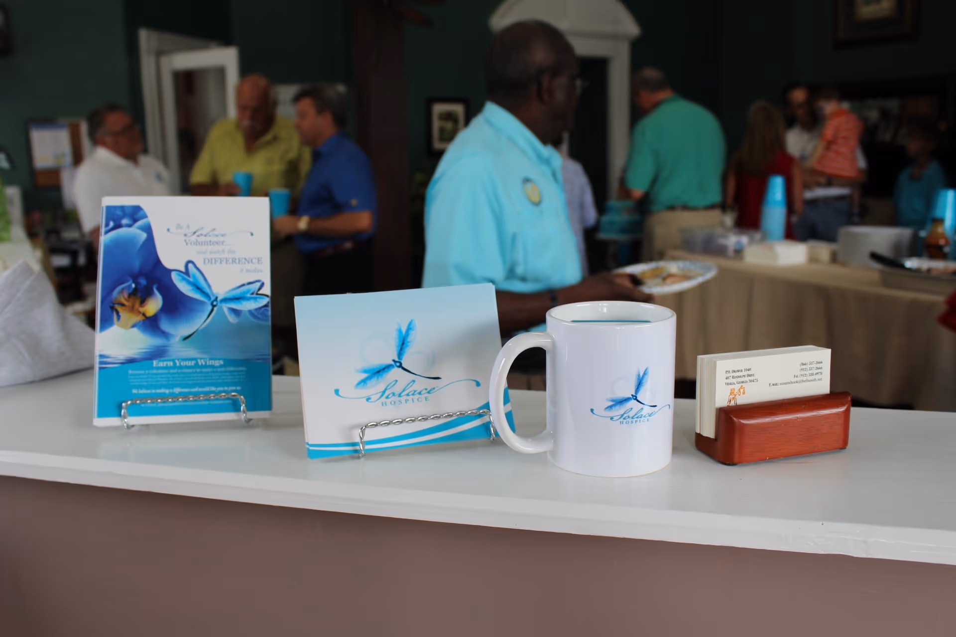 Promotional table with Solace Hospice mug, brochures, and a busy reception area in the background.