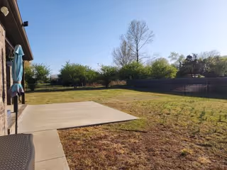 Outdoor patio and grassy yard beside a building, with umbrella, seating, and trees in background.