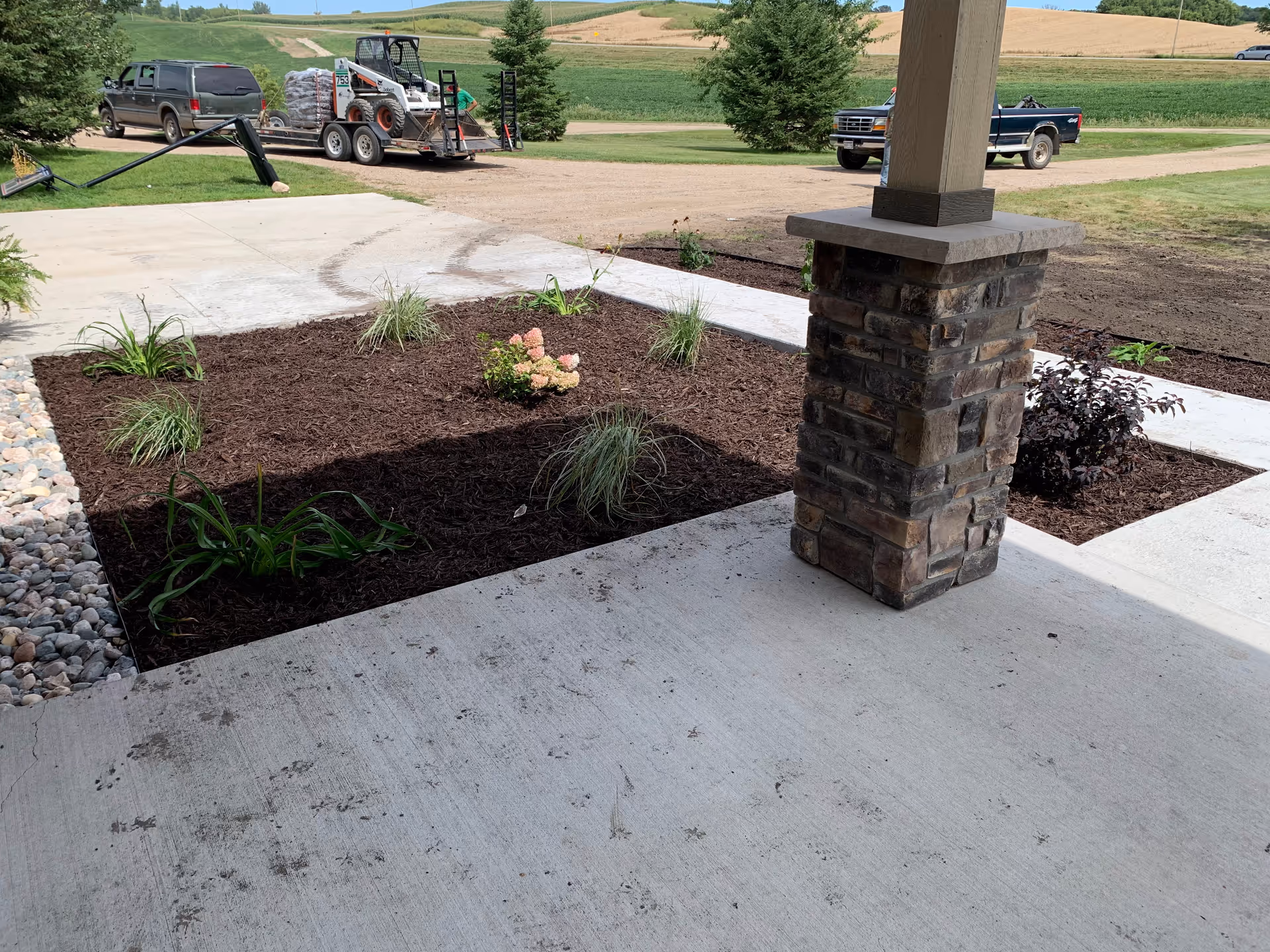 Outdoor entrance area with a stone column, mulch beds, and a concrete walkway.