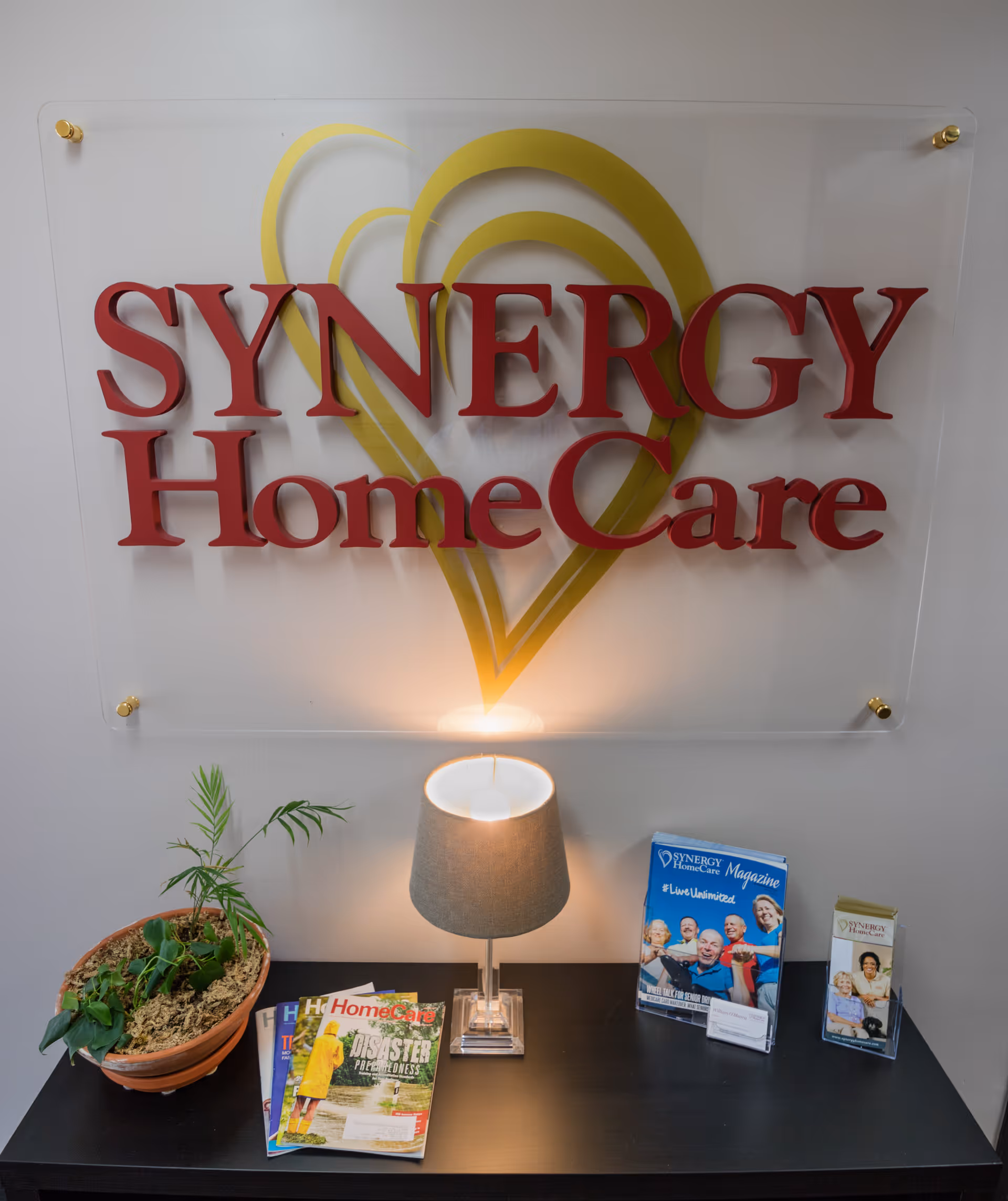 Reception area with Synergy HomeCare sign, lamp, magazines, and a potted plant on a black table.
