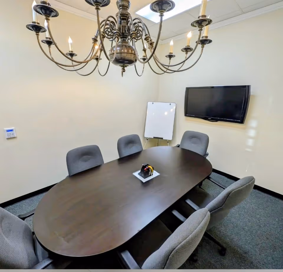 Oval dark wood conference table with gray chairs, chandelier, whiteboard, and wall-mounted TV.