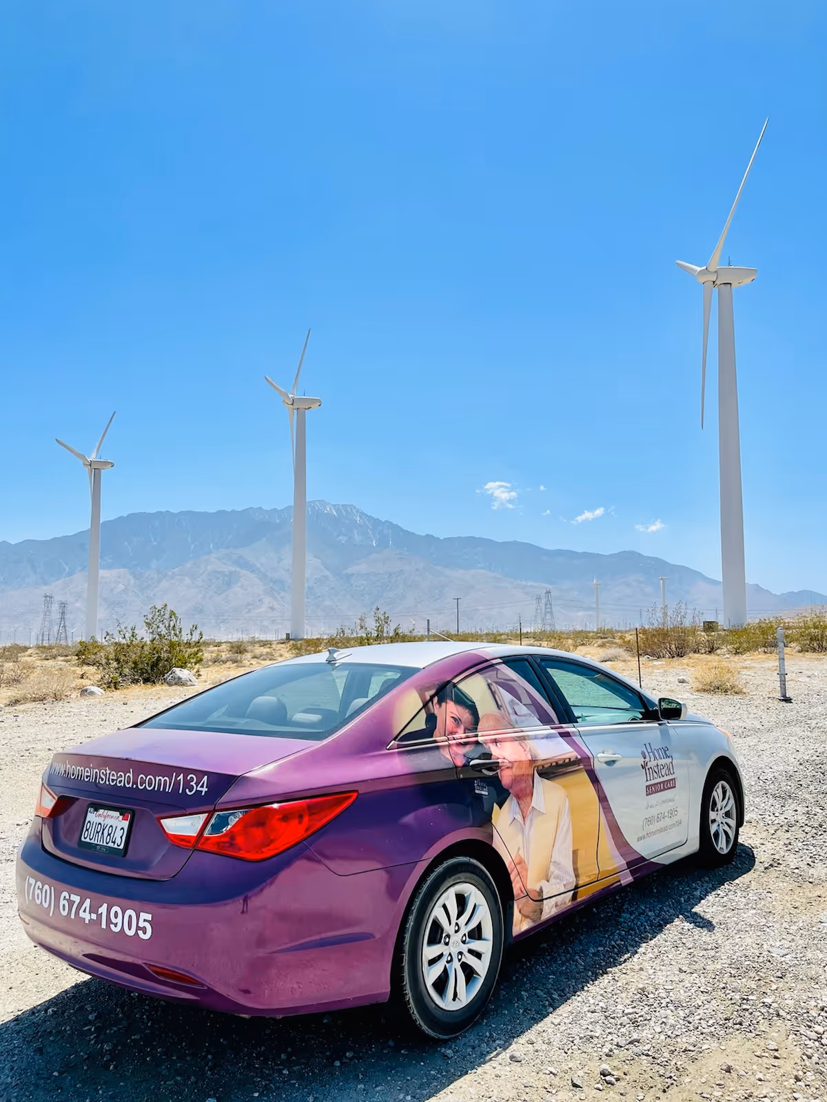 Purple Home Instead car parked on gravel with wind turbines and desert mountains.