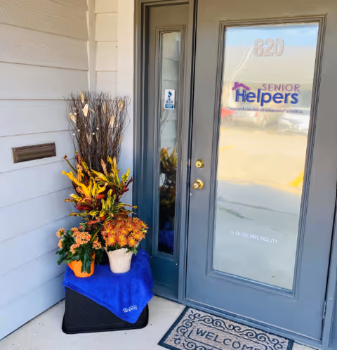 Entrance to Senior Helpers facility with glass door, welcome mat, and decorative plants.