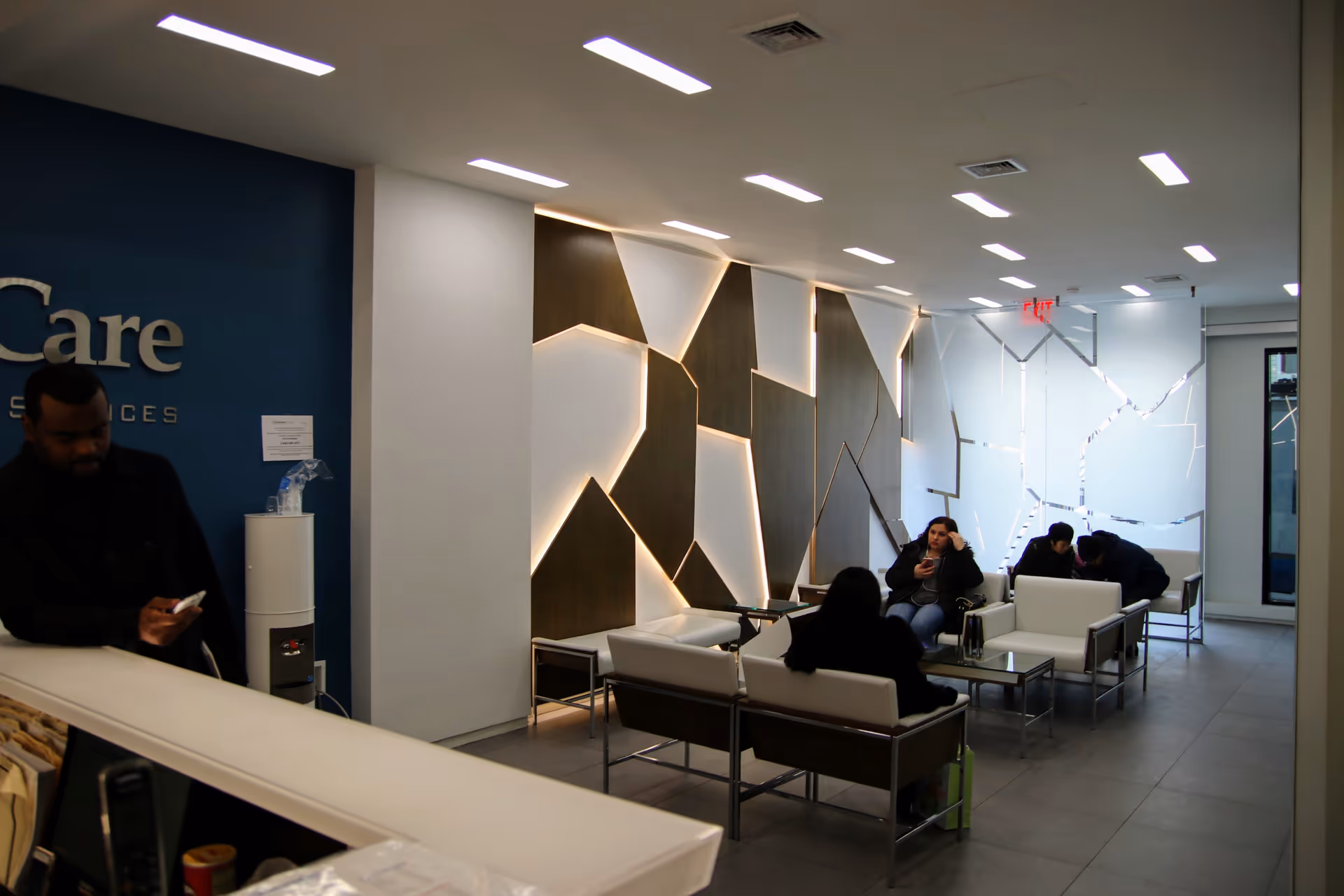 Modern senior care facility lobby with white seating and geometric wall panels; several people seated.