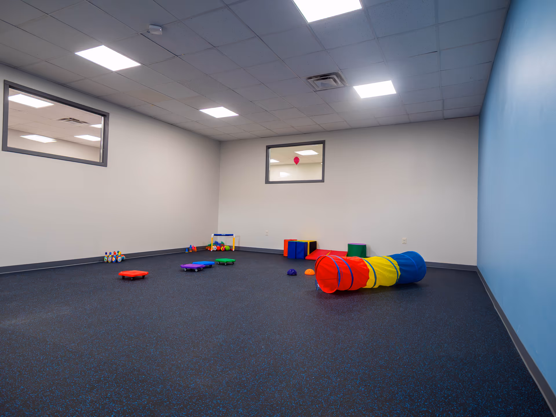 An empty playroom with a colorful tunnel, mats, and small toys in a bright blue-walled room