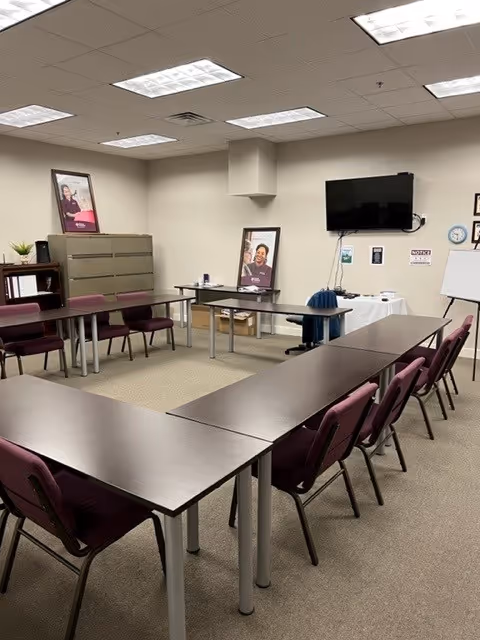 A conference-style meeting room with U-shaped tables, maroon chairs, a wall TV, and posters.