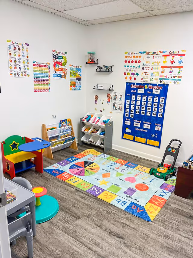 A bright classroom playroom shows alphabet posters, a colorful rug, and toy furniture.