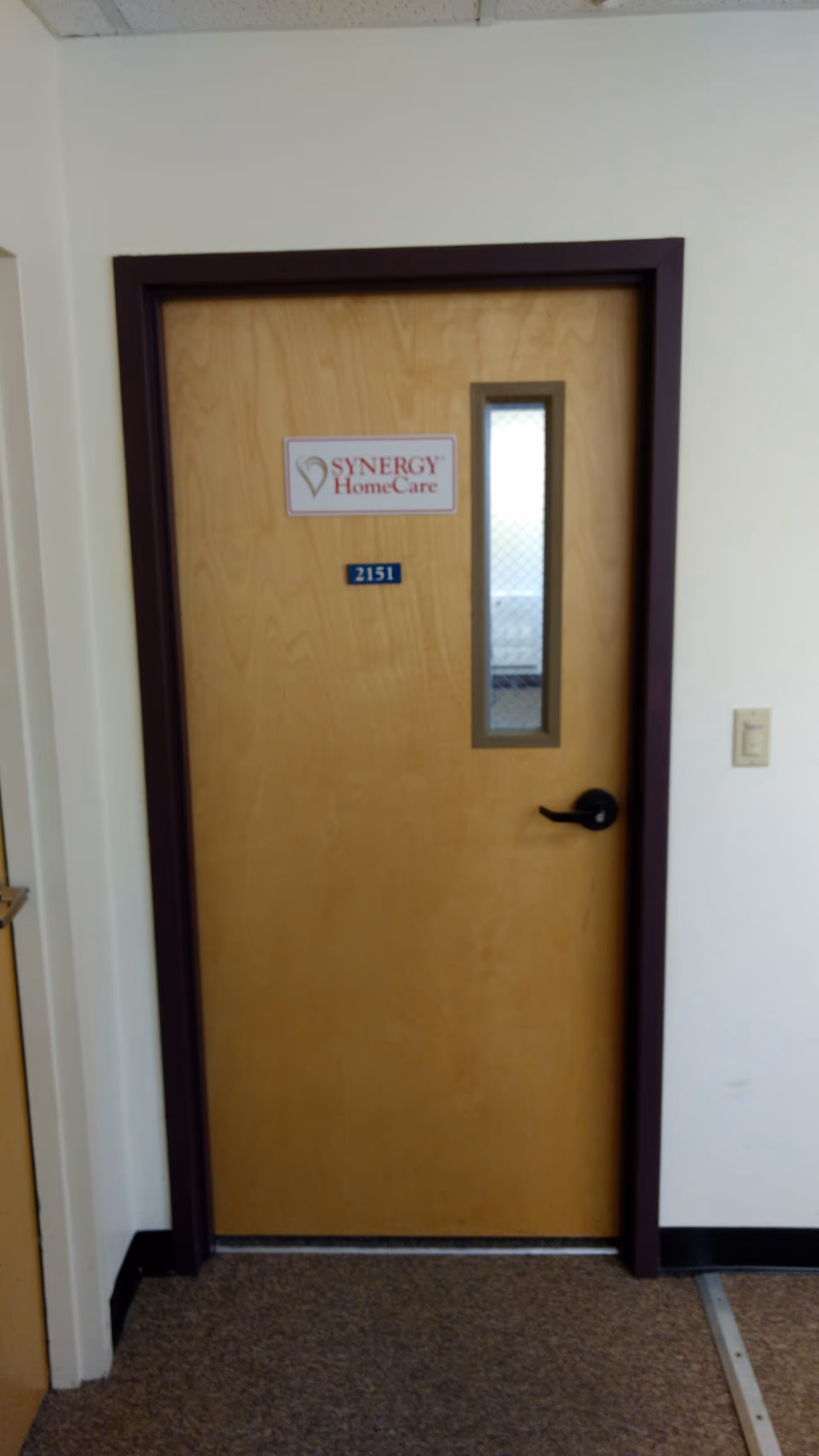Wooden door with small window and SYNERGY HomeCare sign in a hallway.