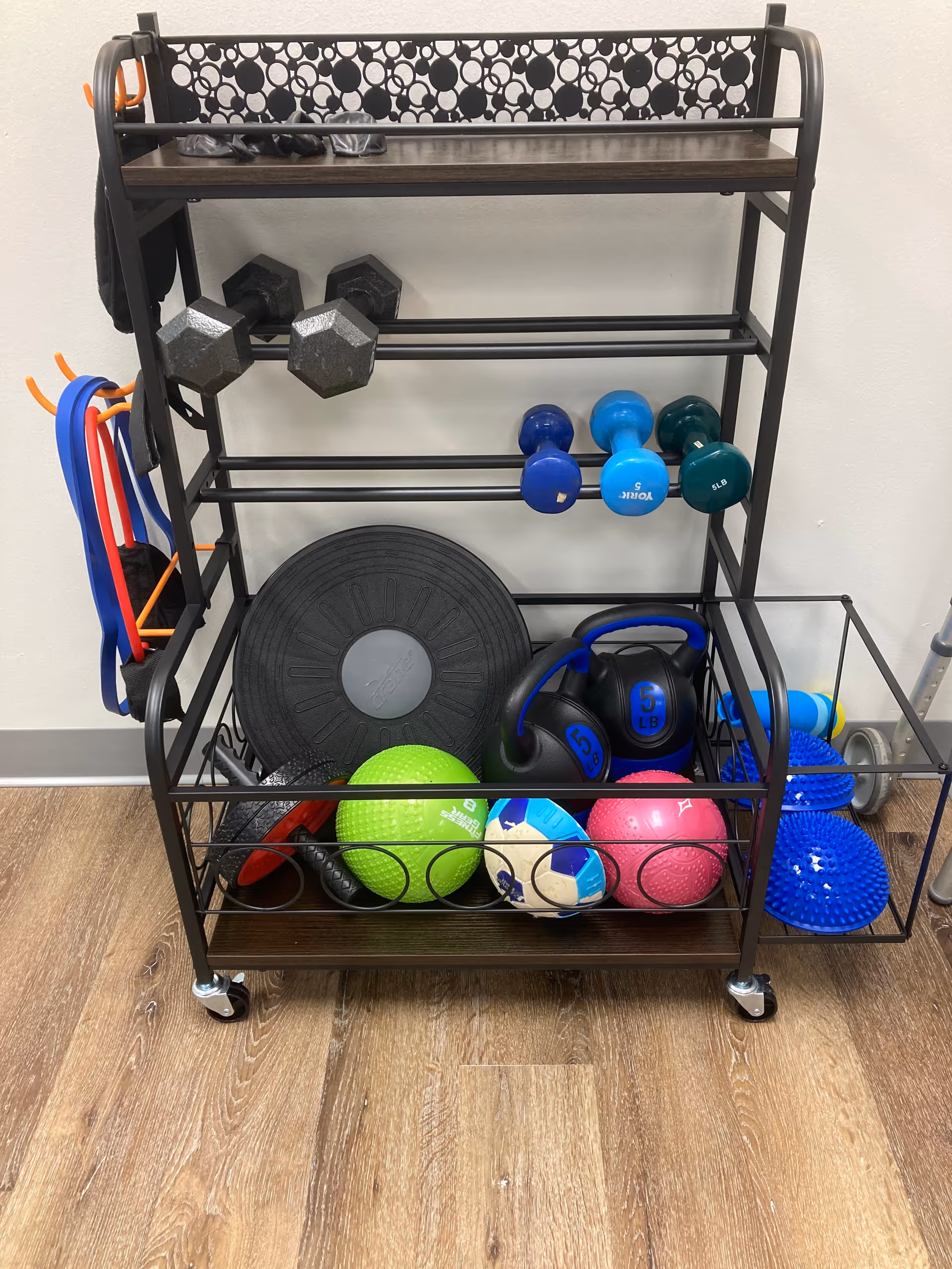 A metal rolling cart in a fitness room holding dumbbells, kettlebells, medicine balls, and bands.